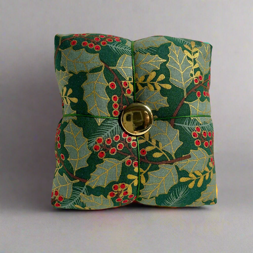 Fabric pincushion with holly leaf pattern and gold button on a gray background