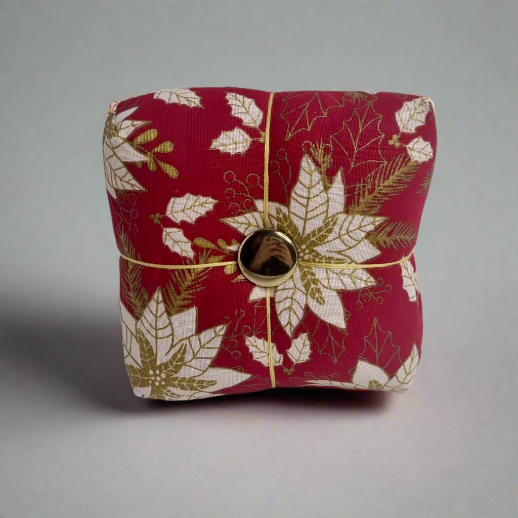 Red fabric pincushion with gold and white leaf pattern and a gold button on a gray background